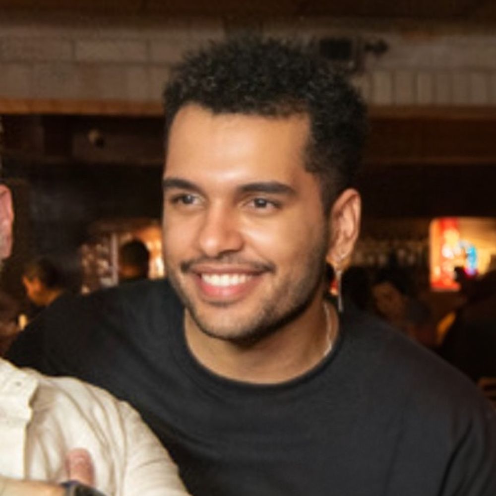 Homem jovem afro-indígena, de pele parda, cabelo curto e cacheado, usando brinco e camiseta preta, sorrindo num ambiente interno de um bar com luz baixa, com pessoas ao fundo desfocadas.