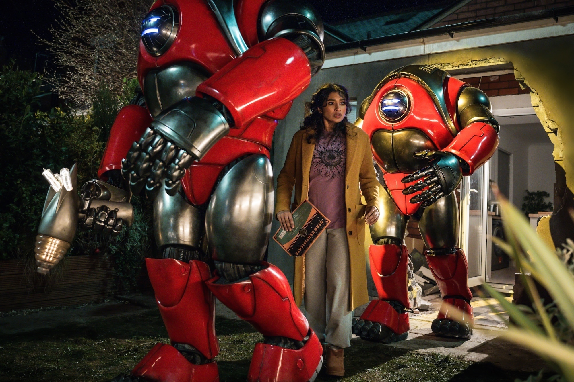 Belinda Chandra (interpretada por Varada Sethu) está cercada por dois grandes robôs vermelhos e prateados do episódio “Robot Revolution” de Doctor Who. Ela parece assustada e confusa, segurando um certificado de nomeação de estrela em meio aos destroços da parede arrombada de sua casa. A cena ocorre à noite, com iluminação dramática e visual que mistura o absurdo cômico dos robôs com o clima tenso da invasão.